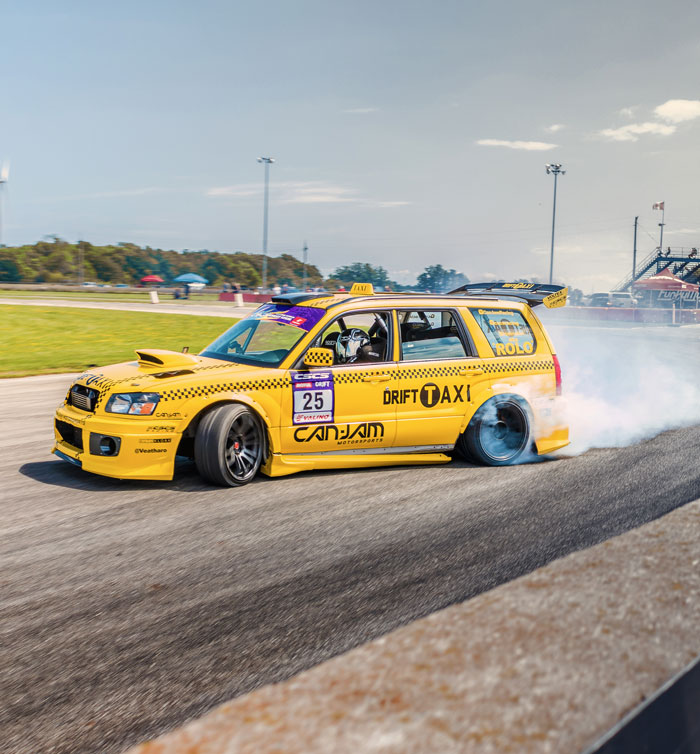 Yellow drift taxi car performing a drift on a motorsport track during a CSCS event, with smoke billowing from the rear tires and a clear blue sky in the background.