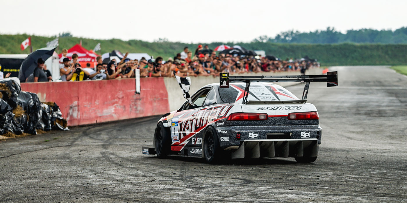 Car drifting on racetrack with checkered flag, surrounded by cheering crowd, showcasing motorsport excitement at CSCS event.