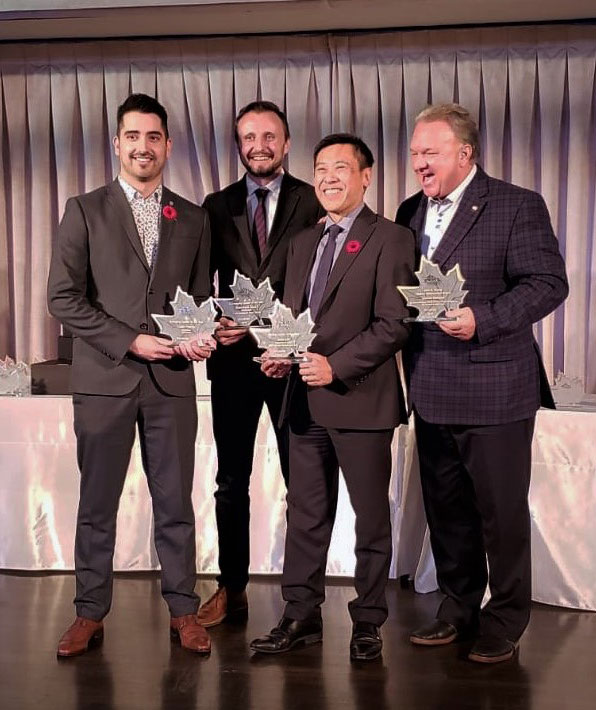Four award recipients smiling and holding glass trophies in a formal setting at the Canadian Printing Awards Gala, with a backdrop of draped fabric and a table displaying additional awards.