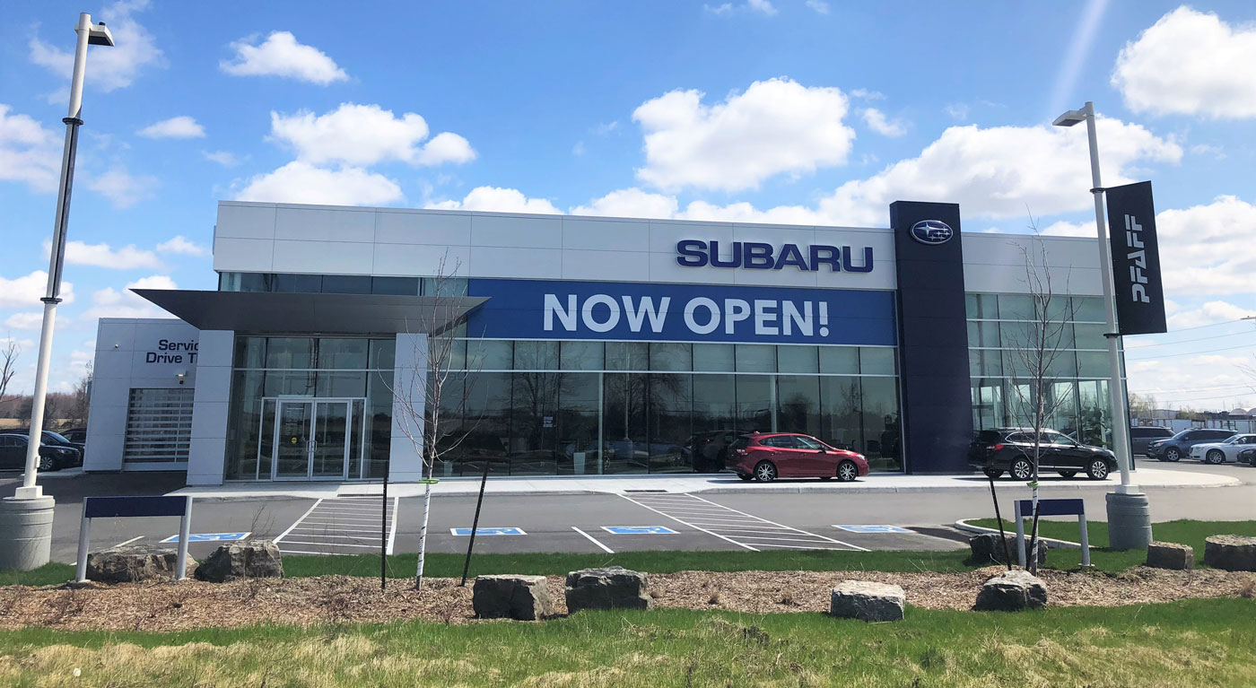 Subaru dealership exterior featuring large "NOW OPEN!" sign, Pfaff branding, and modern architecture with glass windows and landscaped parking area.