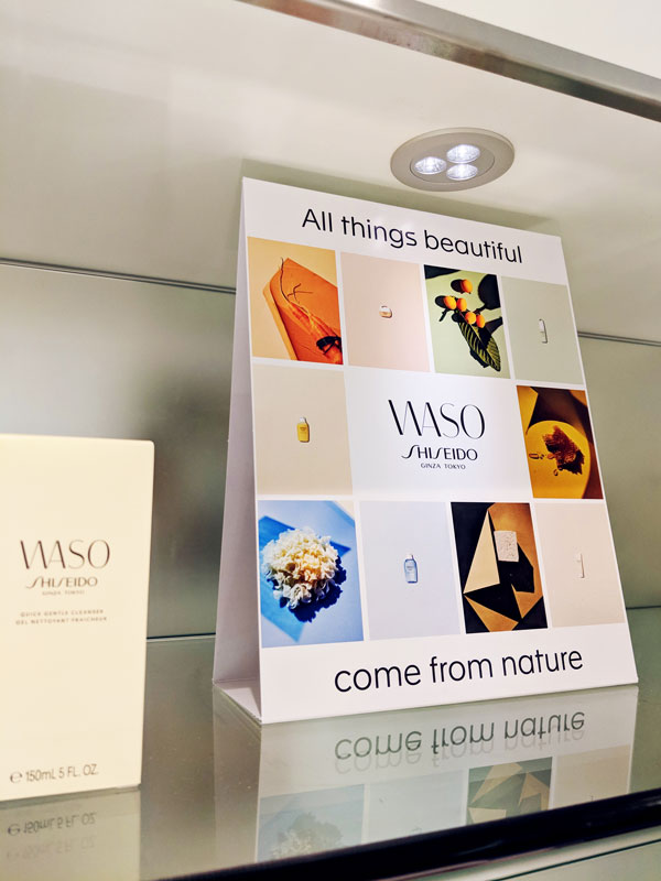 Tent card display featuring WASO by Shiseido with the tagline "All things beautiful" and "come from nature," showcasing product imagery and branding elements on a sleek shelf.