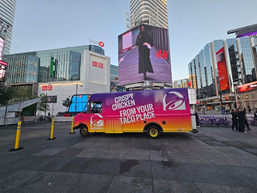 Taco Bell food truck with vibrant branding featuring "Crispy Chicken From Your Taco Place," set in an urban plaza with digital billboards and modern buildings, illustrating fleet branding as a mobile advertising solution.